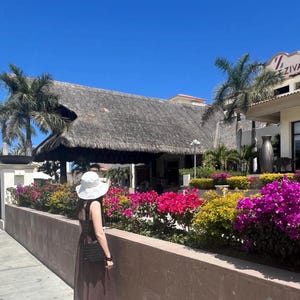 May include: A woman wearing a white sun hat and a brown maxi dress stands in front of a resort building with a thatched roof. The building has a sign that reads "Ziva". There are lush green plants and colorful flowers in the foreground.
