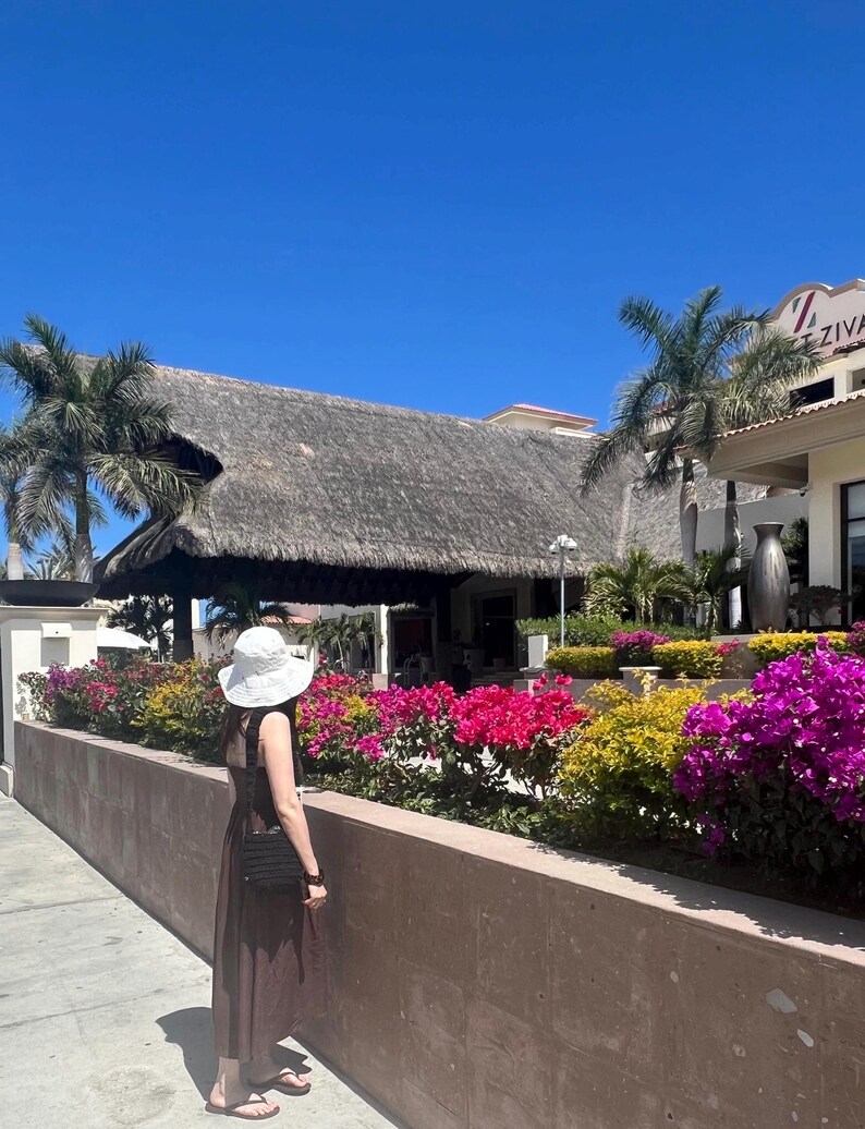 May include: A woman wearing a white sun hat and a brown maxi dress stands in front of a resort building with a thatched roof. The building has a sign that reads "Ziva". There are lush green plants and colorful flowers in the foreground.