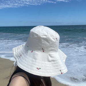 May include: A white bucket hat with red and green embroidered flowers. The hat is worn by a person standing on a sandy beach with ocean waves in the background.