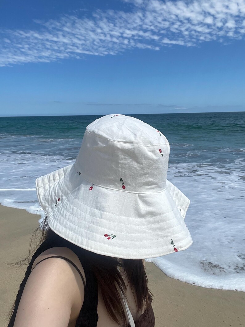 May include: A white bucket hat with red and green embroidered flowers. The hat is worn by a person standing on a sandy beach with ocean waves in the background.