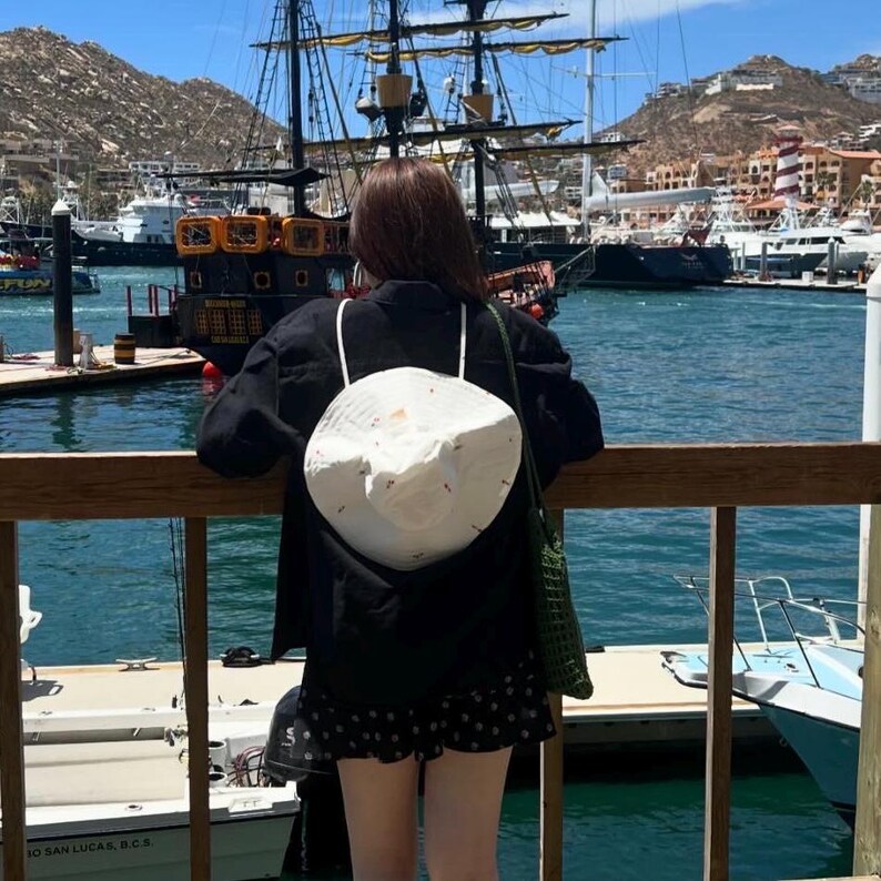 May include: A person wearing a black jacket and a white bucket hat with a floral design stands on a wooden dock overlooking a harbor.  The person is looking out at the water.  There are several boats docked in the harbor, including a large sailing ship.