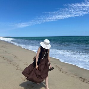 May include: A person wearing a brown maxi dress and a white sun hat walks along a sandy beach with blue water and a clear sky.