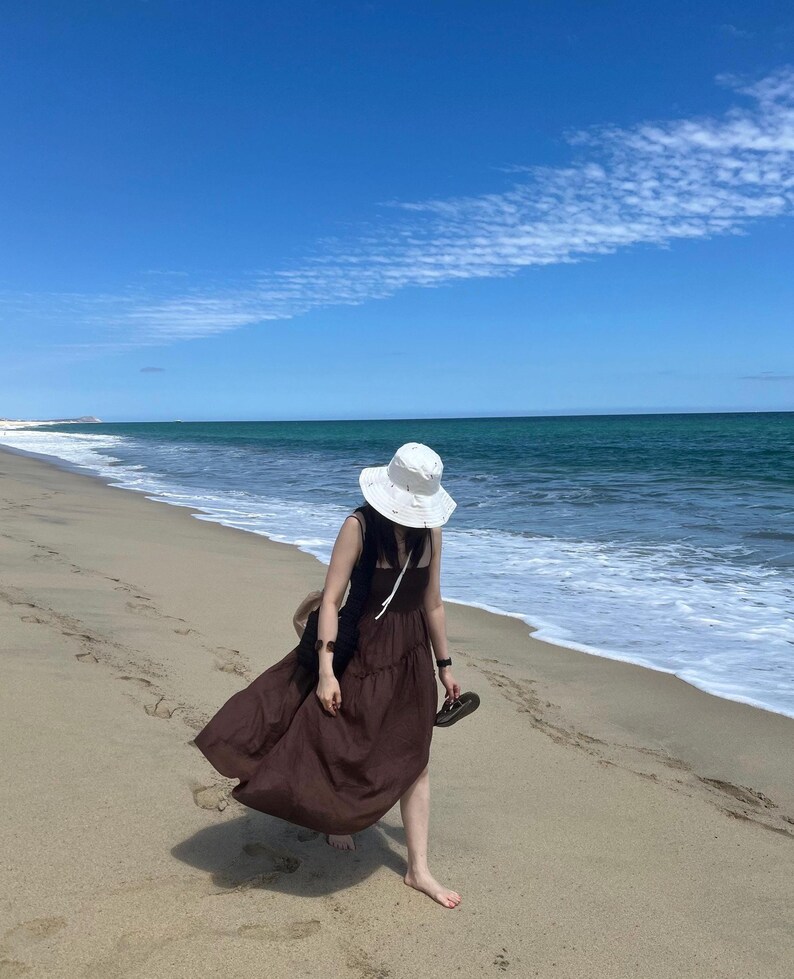 May include: A person wearing a brown maxi dress and a white sun hat walks along a sandy beach with blue water and a clear sky.