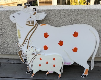 May include: A white cow with orange handprints and a smaller white calf with orange handprints. Both cows are standing on a wooden surface.