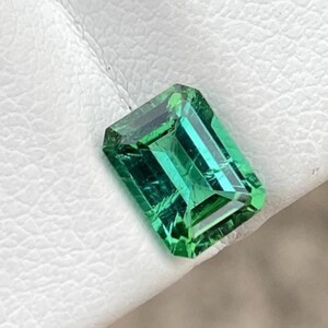 May include: A loose emerald-cut gemstone with a vibrant, saturated green color. The stone features a rectangular shape with faceted edges that create internal light reflections and depth, showcasing a clear, transparent, and polished surface.