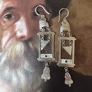May include: Silver earrings with a guillotine design. Each earring has a hook, a helmet-shaped top, a rectangular frame with a triangular blade, and a bell-shaped charm. The earrings are set against a blurred background.