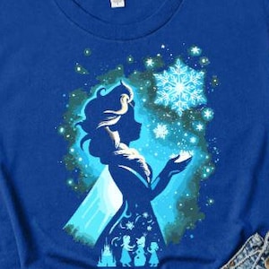 May include: Royal blue t-shirt featuring a silhouette of a woman with a snowflake design. The graphic is in shades of blue and white. The shirt is made of a soft material and has short sleeves.