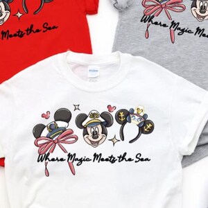 May include: Three short-sleeved t-shirts in red, grey, and white, each featuring a graphic of three mouse-ear headbands and the text Where Magic Meets the Sea. The designs include nautical-themed ears, a captain's hat, and a large pink bow.