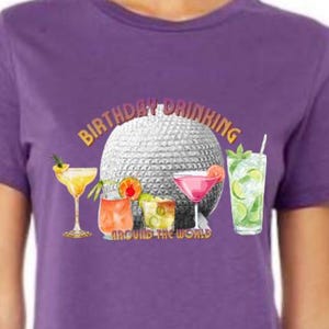 Op de afbeelding: Paarse t-shirt met een afbeelding van een discobal en cocktails. De tekst "BIRTHDAY DRINKING" staat boven de bal, met "AROUND THE WORLD" eronder. Cocktails zijn martini, margarita en mojito, perfect voor een verjaardag.