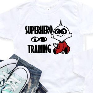 Può includere: Due magliette bianche con la scritta "SUPERHERO IN TRAINING" e una grafica a cartoni animati di un bambino in tuta rossa. Le magliette sono abbinate a jeans blu e un paio di scarpe da ginnastica verde menta e bianche.