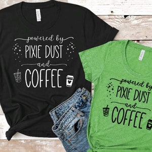 Puede incluir: Dos camisetas con el texto "powered by pixie dust and coffee" impreso en ellas. Una camiseta es negra y la otra es verde.