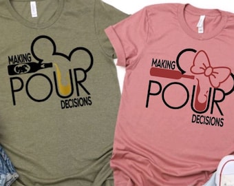May include: Two t-shirts featuring a graphic design of a Mickey Mouse head with a bottle pouring into the ear. The text on the shirts reads "Making Pour Decisions". One shirt is olive green and the other is pink.