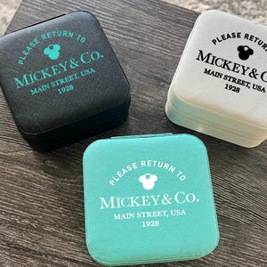 Puede incluir: Tres estuches cuadrados de viaje para joyas en negro, blanco y verde azulado. Cada estuche está impreso con "PLEASE RETURN TO MICKEY & Co. MAIN STREET, USA 1928" en un color contrastante. Los estuches están cerrados y parecen estar hechos de un material texturizado.