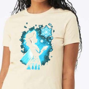 May include: Cream-colored t-shirt featuring a blue silhouette of a character with a snowflake design. The graphic includes a snowflake and other icy elements. The shirt is a short-sleeved crew neck.