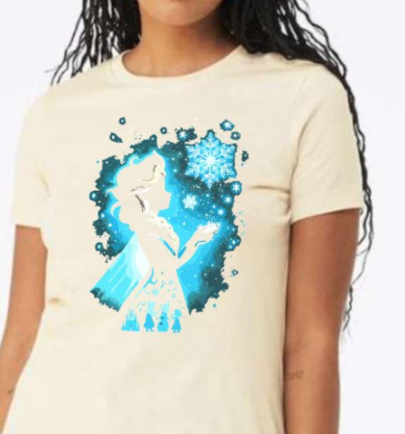 May include: Cream-colored t-shirt featuring a blue silhouette of a character with a snowflake design. The graphic includes a snowflake and other icy elements. The shirt is a short-sleeved crew neck.