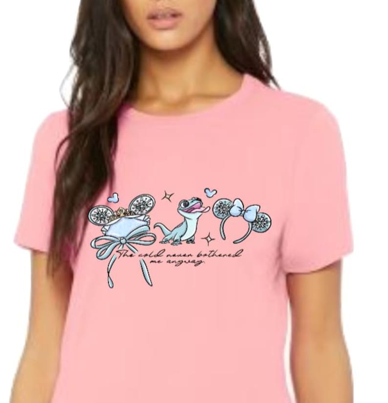 以下が含まれることがあります： 淡いピンク色の半袖Tシャツ。青いトカゲ、2組のネズミの耳、そして「The cold never bothered me anyway.」というテキストのグラフィックがプリントされています。デザインには雪の結晶の模様、王冠、耳のヘッドバンドに付いたリボンが含まれています。