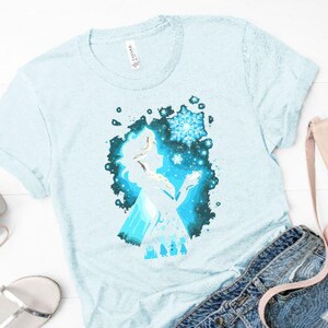 May include: Light blue t-shirt featuring a silhouette of a character with a snowflake design. The graphic is in shades of blue and white. The shirt is paired with denim shorts, a purse, headphones, and sandals.