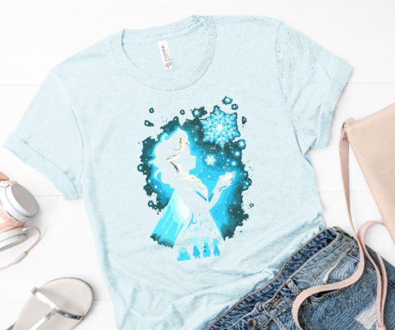 May include: Light blue t-shirt featuring a silhouette of a character with a snowflake design. The graphic is in shades of blue and white. The shirt is paired with denim shorts, a purse, headphones, and sandals.
