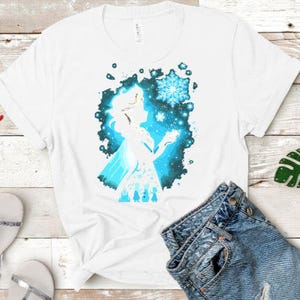 May include: White t-shirt featuring a silhouette of a woman with a snowflake design. The graphic is in shades of blue and white, with a splash effect background. The shirt is paired with denim shorts.