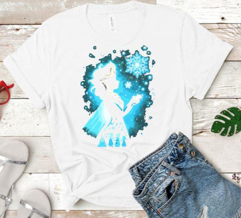 May include: White t-shirt featuring a silhouette of a woman with a snowflake design. The graphic is in shades of blue and white, with a splash effect background. The shirt is paired with denim shorts.