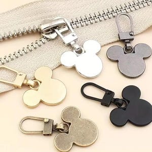 Może przedstawiać: Pięć suwaków do zamków błyskawicznych z motywem głowy Myszki Miki. Suwaki są w kolorze srebrnym, złotym, czarnym i brązowym.