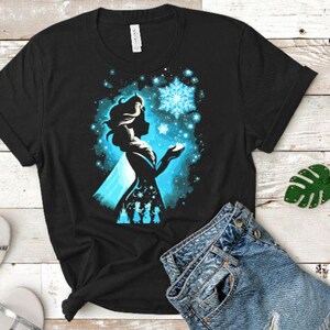 May include: Black t-shirt featuring a silhouette of a woman with a blue and white snowflake design. The design includes a silhouette of three figures at the bottom. The shirt is made of a soft material.