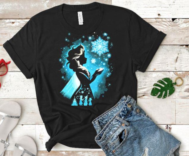 May include: Black t-shirt featuring a silhouette of a woman with a blue and white snowflake design. The design includes a silhouette of three figures at the bottom. The shirt is made of a soft material.