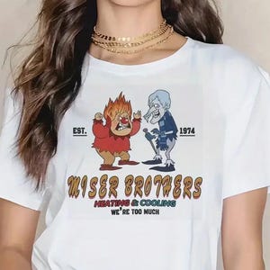 May include: White t-shirt featuring a graphic of two cartoon characters, one with flames for hair and the other with a blue coat. The text "Miser Brothers Heating & Cooling" and "We're Too Much" is printed below the characters. The shirt also has "EST. 1974" printed above the characters.