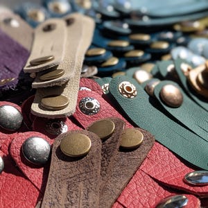 May include: A collection of multi-coloured leather wristbands featuring textured surfaces and metallic snap button closures in brass and silver tones. The assortment includes various colours such as red, green, purple, yellow, and beige leather straps.