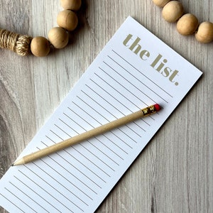 May include: A white notepad with gold lined paper and the text "the list." printed in gold. A wooden pencil with a red eraser is lying on top of the notepad.