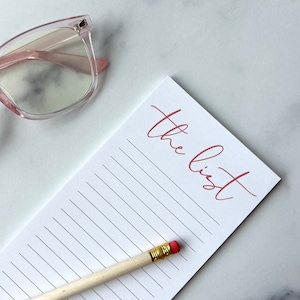 May include: A white notepad with the words "the list" written in pink cursive. A pencil with a pink eraser is lying on the notepad.
