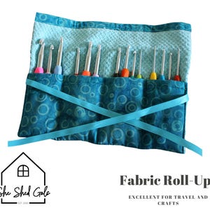 May include: A blue fabric roll-up with a circular pattern, holding several crochet hooks with colorful handles. The roll-up is secured with a blue ribbon. Text on the image reads "Fabric Roll-Ups" and "Excellent for Travel and Crafts."