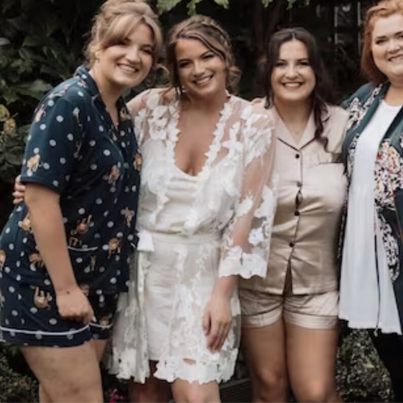 May include: Four women in a group photo, wearing pajamas. The woman in the center is wearing a white lace robe. The women on either side of her are wearing dark blue and light brown pajamas. The woman on the far right is wearing a green and black patterned jacket.