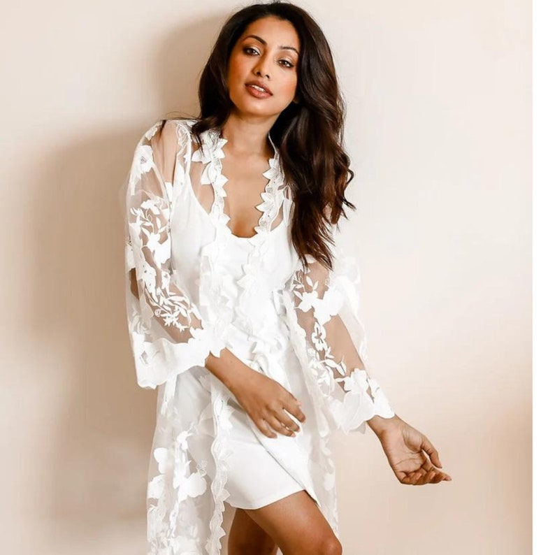 May include: White lace robe with floral embroidery. The robe has long sleeves and a V-neckline. The model is wearing a white slip dress underneath.
