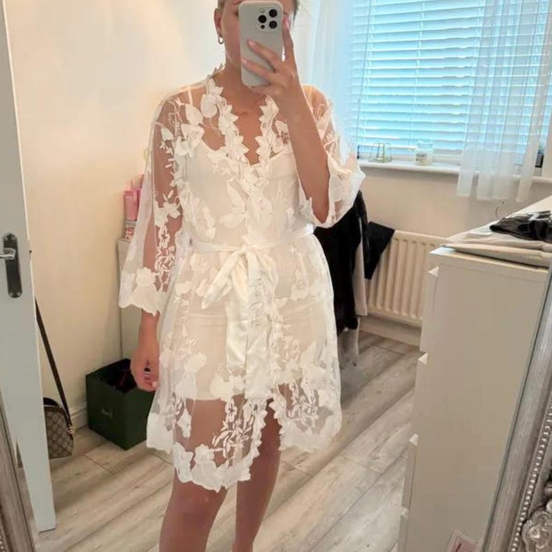May include: A white lace robe with floral embroidery and a satin tie belt. The robe is sheer and has a delicate, feminine look.