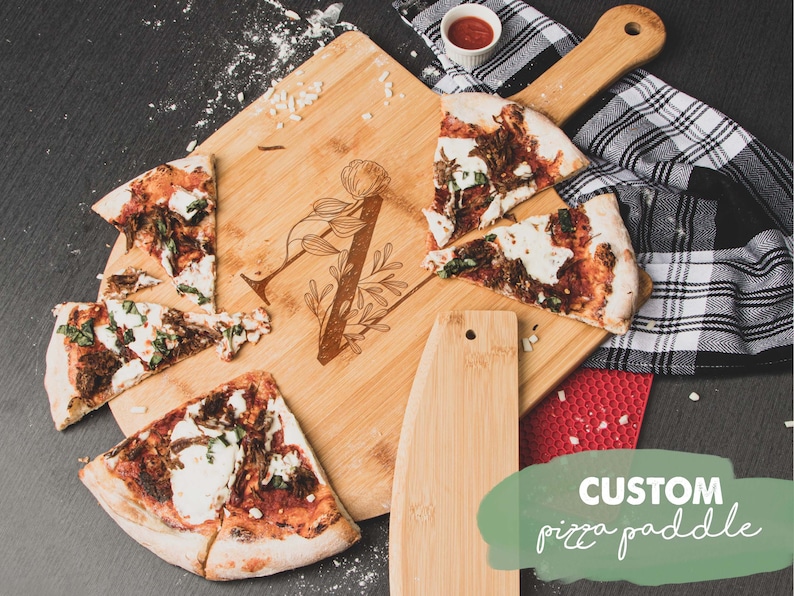 May include: A wooden pizza paddle with a handle and a built-in pizza cutter. The paddle has a floral design and the text "CUSTOM pizza paddle" is written on the bottom right corner of the image.