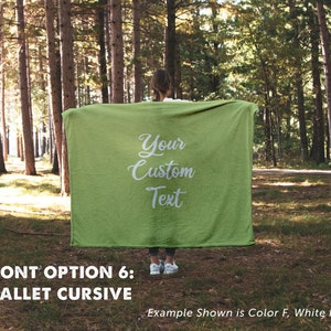 May include: A green fleece blanket with white cursive text that reads "Your Custom Text". The blanket is held up by a person in a forest setting. The text "FONT OPTION 6: BALLET CURSIVE" is below the image. The text "Example Shown is Color F, White Font" is below the text.