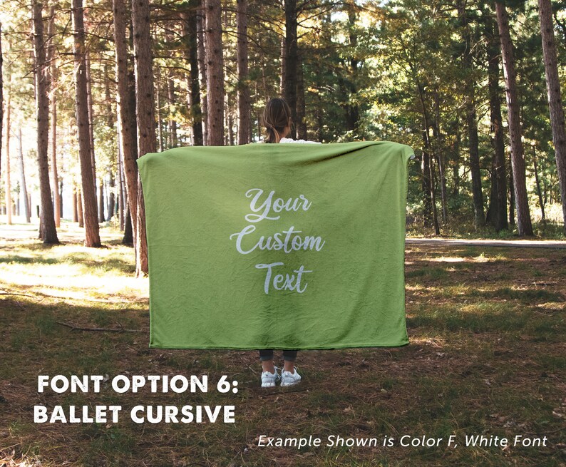 May include: A green fleece blanket with white cursive text that reads "Your Custom Text". The blanket is held up by a person in a forest setting. The text "FONT OPTION 6: BALLET CURSIVE" is below the image. The text "Example Shown is Color F, White Font" is below the text.