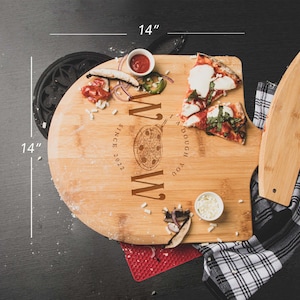 May include: A 14-inch round bamboo pizza cutting board with a built-in pizza cutter. The board has a personalized message that reads "You Dough My " with a pizza slice graphic and "Since 2022" below. The board is shown with two slices of pizza, a small bowl of cheese, and a small bowl of red sauce.