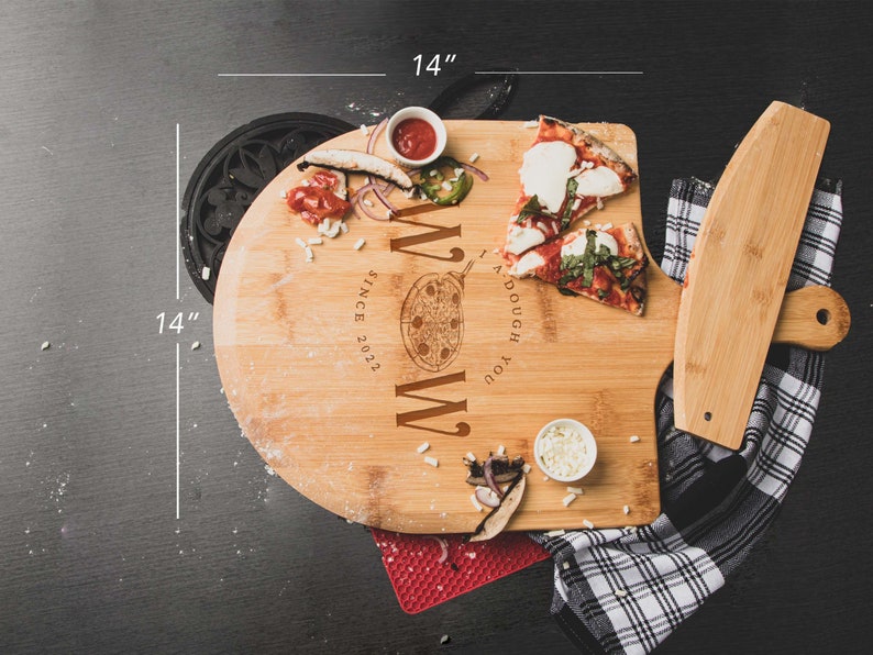 May include: A 14-inch round bamboo pizza cutting board with a built-in pizza cutter. The board has a personalized message that reads "You Dough My " with a pizza slice graphic and "Since 2022" below. The board is shown with two slices of pizza, a small bowl of cheese, and a small bowl of red sauce.