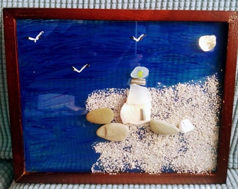 May include: A framed artwork depicting a coastal scene. A lighthouse made of sea glass and shells stands on a sandy shore, with a blue ocean and seagulls overhead. The frame is dark brown.