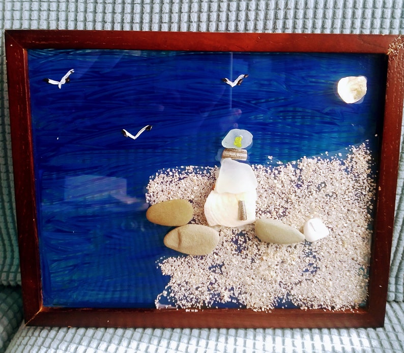 May include: A framed artwork showing a coastal scene. A lighthouse made of sea glass and shells stands on a sandy shore, with a blue ocean and seagulls overhead. The frame is dark brown.