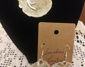 Può includere: Un set di collana e orecchini in conchiglia. La collana presenta un grande ciondolo a forma di conchiglia con un designo a ramo argentato. Gli orecchini sono realizzati con conchiglie più piccole con ganci argentati. L'etichetta recita "Seashell Equation".