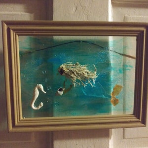May include: A framed diorama depicting an underwater scene. It includes a seahorse, a shell, and dried plant matter, set against a blue and green painted backdrop. The frame is made of light brown wood, and the scene is enclosed in glass.