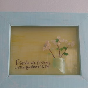 May include: A framed piece of art with a yellow background and a quote that reads "Friends are flowers in the garden of life". The art features a bouquet of white flowers made from sea glass in a green glass vase.