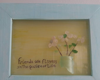 Può includere: Un'opera d'arte incorniciata con uno sfondo giallo e una citazione che recita "Gli amici sono fiori nel giardino della vita". L'opera presenta un bouquet di fiori bianchi realizzati in vetro di mare in un vaso di vetro verde.