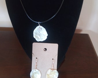 Può includere: Un set di gioielli in conchiglia con una collana con un ciondolo a forma di conchiglia grande e un paio di orecchini con due conchiglie più piccole ciascuno. La collana è su un cordoncino nero e gli orecchini hanno ganci dorati.