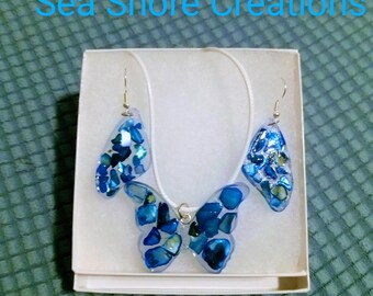 Può includere: Un set di collana e orecchini a forma di farfalla blu su sfondo bianco. I gioielli sono realizzati con conchiglie blu e iridescenti.