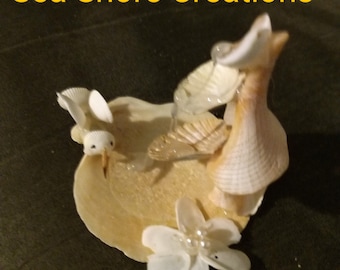 Può includere: Una scultura di un uccello realizzata con conchiglie marine con una cascata che scende lungo la schiena. La scultura è decorata con fiori bianchi e perle. La scultura è realizzata con conchiglie marine naturali ed è un'opera d'arte unica e bellissima.