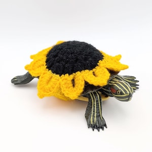 May include: A realistic turtle figurine wearing a crocheted sunflower cover on its shell. The sunflower features a dark black center with bright yellow petals. The turtle has a dark green body with yellow stripes and a distinct red patch near its eye.